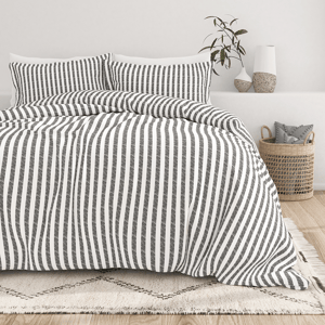 A bed with a black and white vertical striped duvet and pillows is set in a minimalist bedroom. A woven basket and decorative vases complete the decor.
