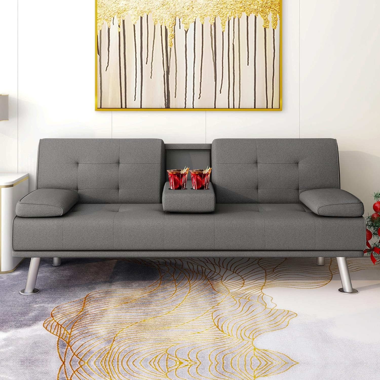 Gray futon sofa bed with a split back design, featuring two removable armrests and a center fold-down console with cup holders. The sofa stands on metallic legs and is placed on a decorative rug with a gold line pattern.