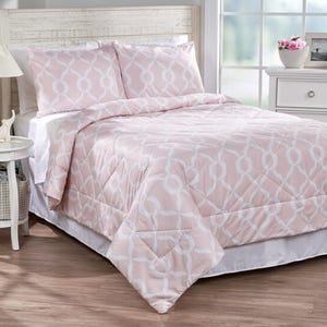 A bed is adorned with a pink comforter and pillowcases featuring a white geometric pattern, set in a bright, cozy bedroom with a white headboard and nightstand.