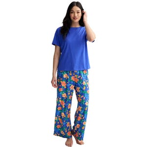 A woman models a two-piece pajama set featuring a solid blue short-sleeve top paired with loose-fitting pants that have a vibrant floral pattern in orange and pink hues on a blue background.