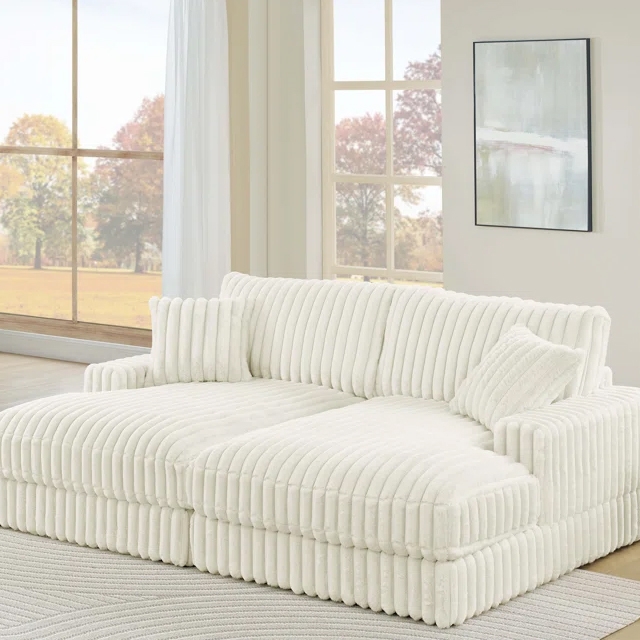 A large, plush, white sectional sofa with ribbed upholstery is positioned in a bright room with large windows and minimalist decor, including a light abstract painting on the wall.