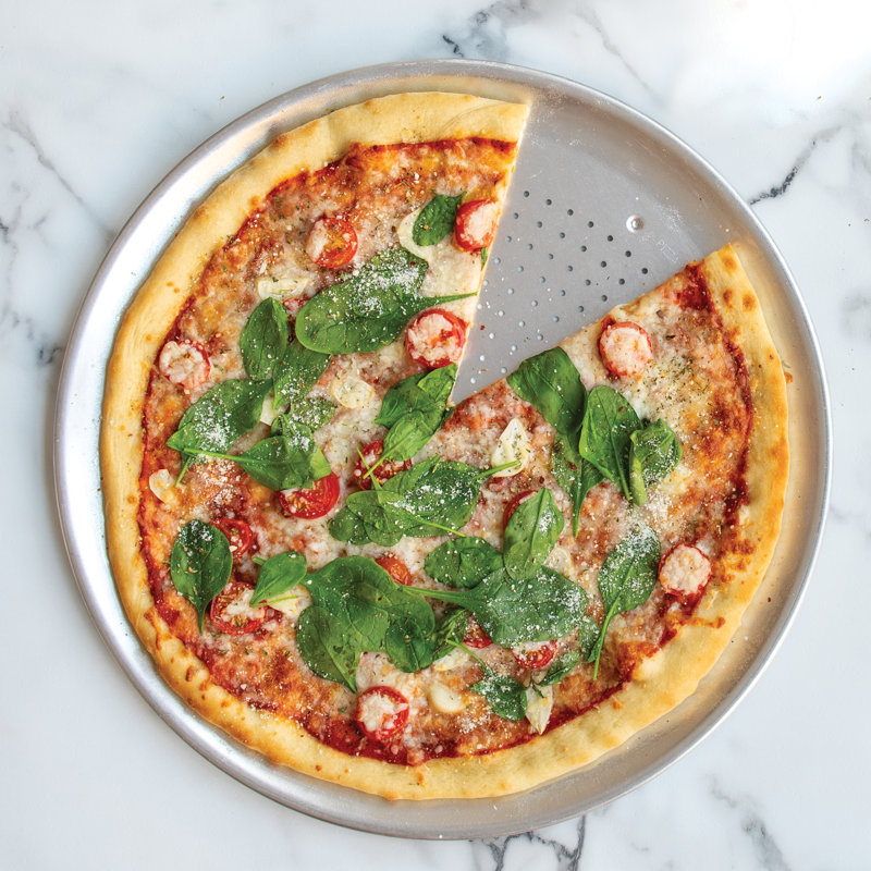 A round pizza topped with spinach, sliced tomatoes, cheese, and a sprinkle of herbs and parmesan, served on a metal pizza tray, with one slice partially moved.