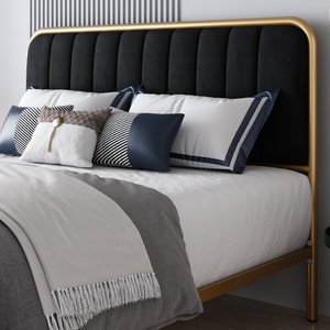 A bed with a black upholstered headboard featuring a gold frame, adorned with white and navy pillows, including one with a distinctive geometric pattern, and a gray blanket with fringe detailing.