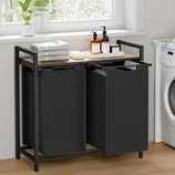 A two-compartment black laundry hamper with a metal frame, featuring a wooden top used to hold folded towels and toiletry items. It is positioned next to a washing machine in a laundry room.