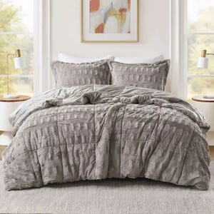 A gray faux fur queen comforter set includes a plush comforter and matching pillow shams, featuring a subtle checkered texture. The bed is styled in a cozy, modern bedroom with abstract wall art and minimalist bedside lamps.