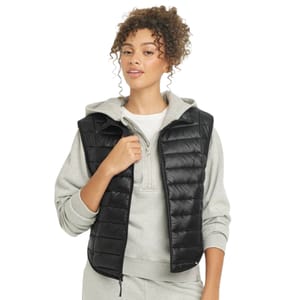 The women's full-zip puffer vest is black with a quilted design, sleeveless style, and front zipper. It is worn over a light grey hoodie for a casual layered look.