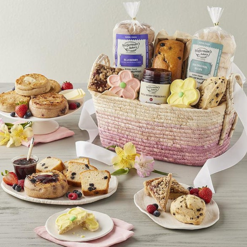 A gift basket includes blueberry and traditional English muffins, a lemon poppy seed loaf, fruit preserves, flower-shaped cookies, and assorted pastries, displayed with fresh berries and flowers.