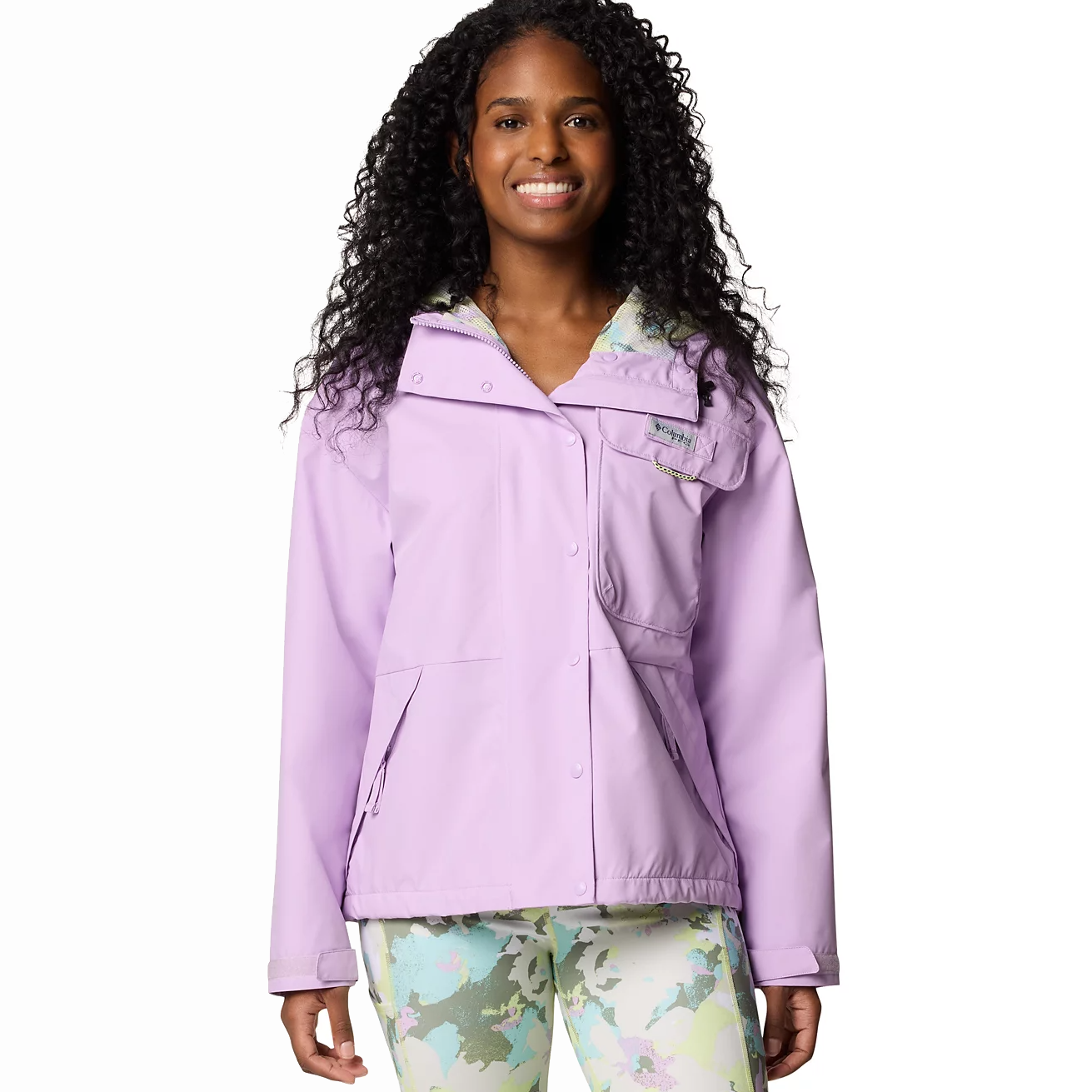 A woman is wearing a Columbia Hooded Rain Jacket in light purple with a front pocket and snap closures, complemented by colorful patterned pants.