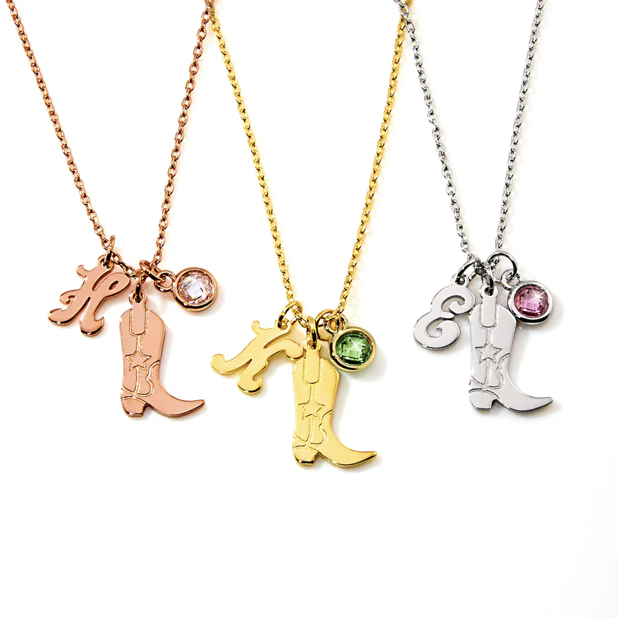 Three necklaces feature charms: a cowboy boot, an initial letter, and a gemstone. One is rose gold with a clear gem, one is yellow gold with a green gem, and one is silver with a pink gem.