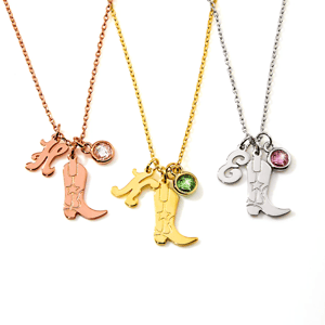 Three necklaces feature charms: a cowboy boot, an initial letter, and a gemstone. One is rose gold with a clear gem, one is yellow gold with a green gem, and one is silver with a pink gem.