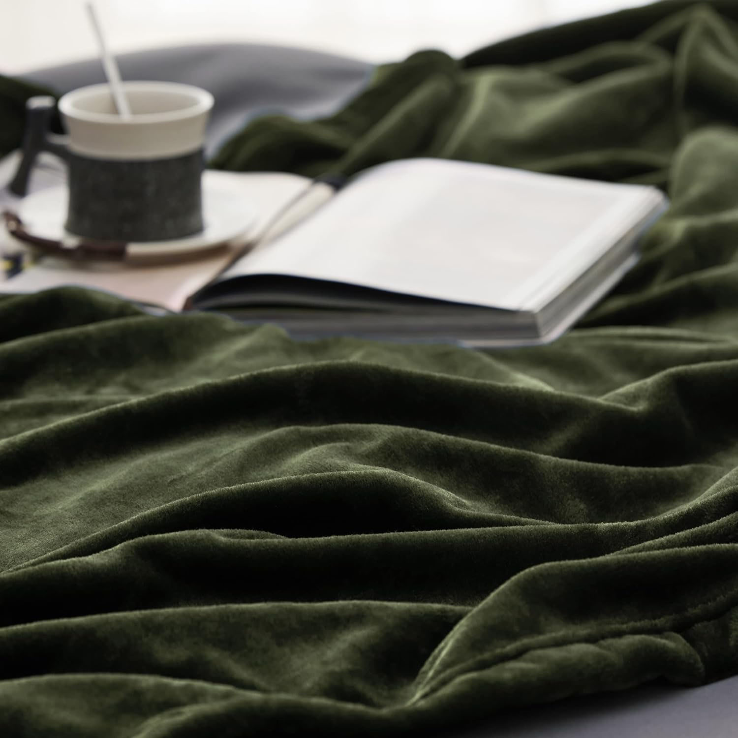 A soft green blanket is draped beside an open book and a coffee cup on a saucer, creating a cozy setting.