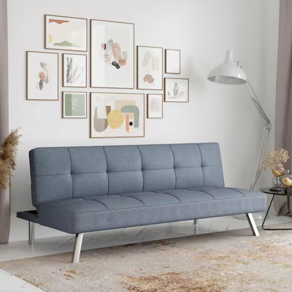Modern living room features a blue futon-style sofa with chrome legs, artistic wall gallery with abstract prints, a tall white floor lamp, and a round side table with decorative elements, set on a neutral-patterned rug.