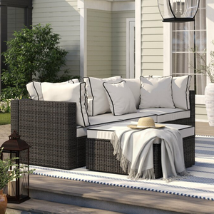 A wicker outdoor patio set with cream cushions, accompanied by striped pillows, a throw blanket, and a sun hat on the ottoman.