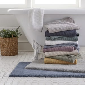 A stack of neatly folded towels in various colors next to a bathtub with a plant in a wicker basket and bath mats on the floor.