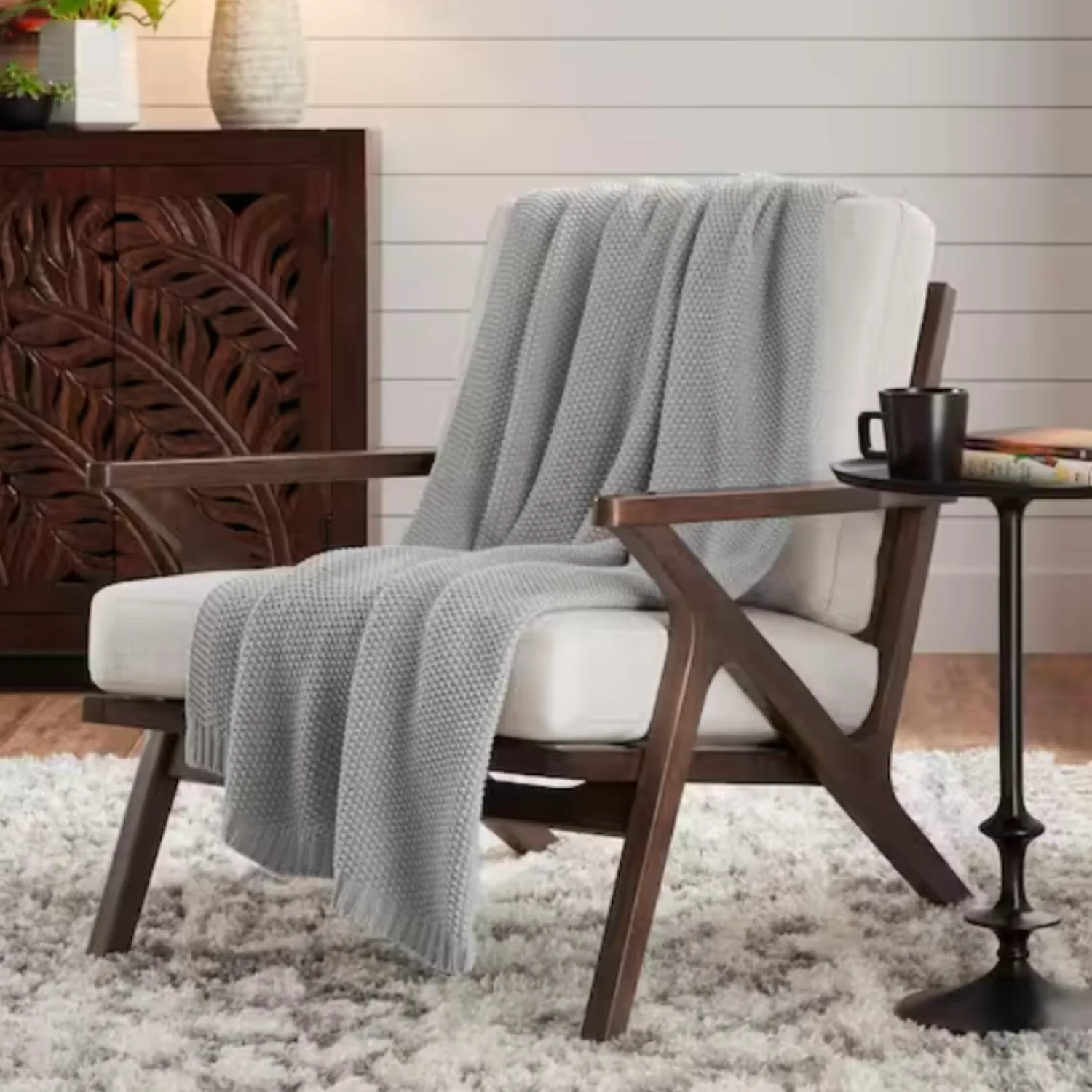 A light gray knit throw blanket is draped over a cushioned wooden chair, which is placed on a thick, white shag rug.
