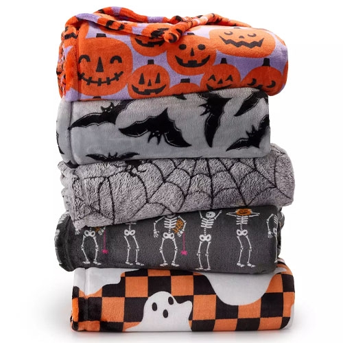 A stack of Halloween-themed blankets featuring designs with jack-o'-lanterns, bats, spiderwebs, skeletons, and ghosts on a checkered background.