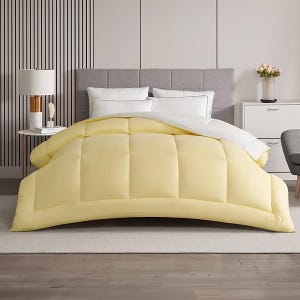 A bedroom setup features a bed with a light yellow duvet and white pillows. The room includes a gray upholstered headboard, a modern bedside lamp, a small side table with books, and a white dresser with a plant.