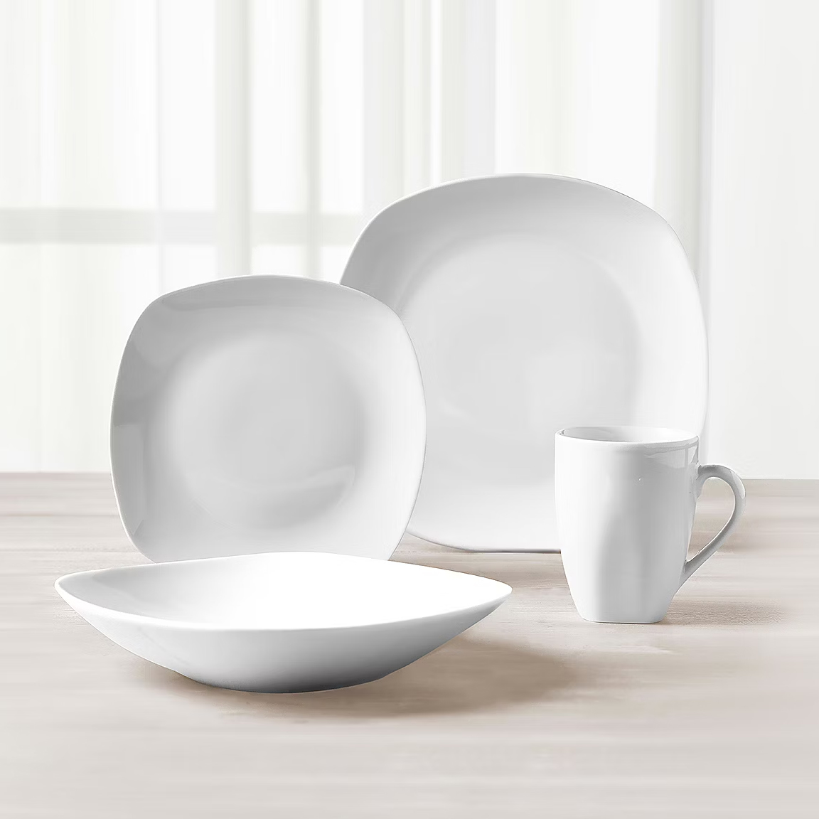 White dinnerware set with two plates, a bowl, and a mug on a wooden table with sheer curtains in the background.