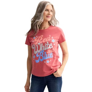 A woman wears a pink T-shirt with the phrase \“Red, White and Blue\“ in bold, colorful letters surrounded by stars, paired with dark jeans.