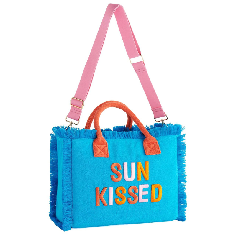 A blue tote bag with fringe details, pink and orange fabric handles, and a pink adjustable shoulder strap. The front features the words \“SUN KISSED\“ in large colorful letters.