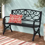 Black metal bench with ornate backrest design, featuring a red and white geometric patterned pillow. Placed on a wooden floor, flanked by potted plants and a striped rug.