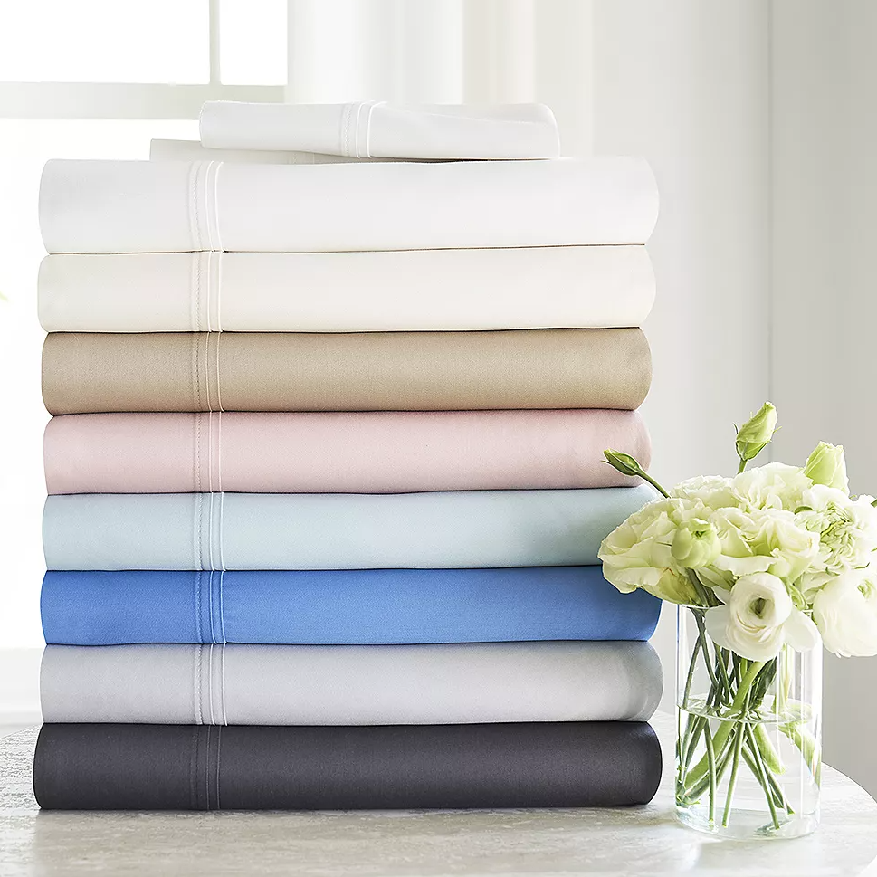 A stack of folded bed sheets in various colors, accompanied by a vase of white flowers.