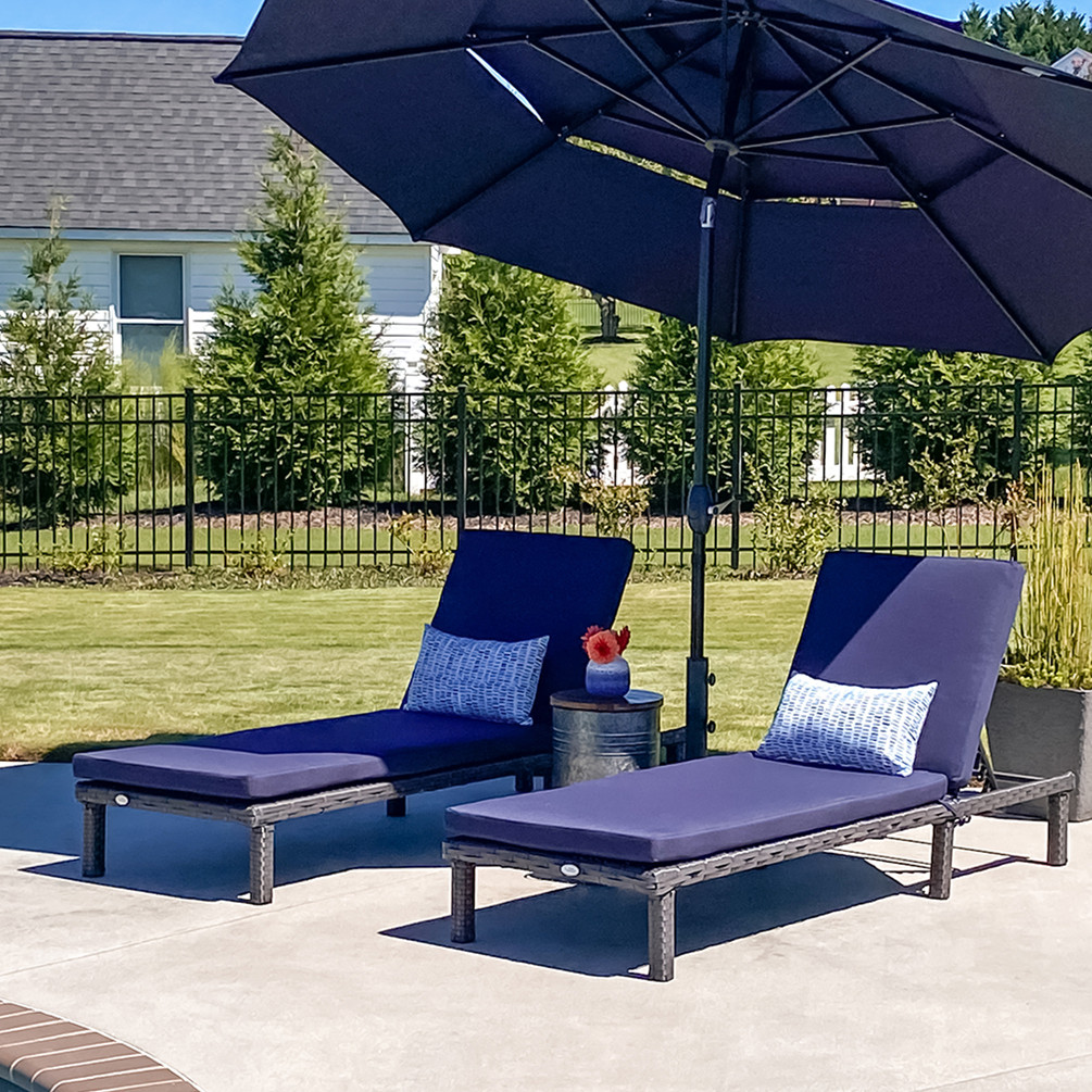 Two outdoor chaise lounge chairs with navy blue cushions and pillows sit under a large navy umbrella, next to a small round side table with a vase and orange flower, placed by a poolside in a backyard setting.