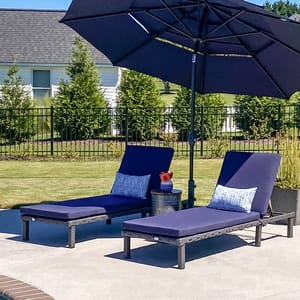 Two outdoor chaise lounge chairs with navy blue cushions and pillows sit under a large navy umbrella, next to a small round side table with a vase and orange flower, placed by a poolside in a backyard setting.
