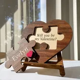 A wooden heart-shaped puzzle with a detachable piece is inscribed with \“Will you be my Valentine?\“ on the heart and \“Evelyn\“ with a red heart on the piece.