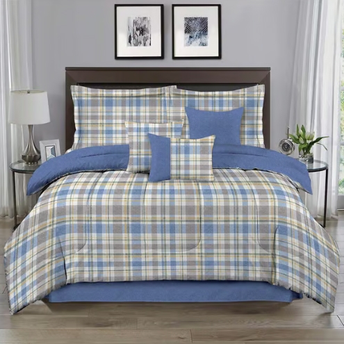 A neatly made bed with a plaid comforter set, featuring blue accents and multiple pillows in plaid and solid blue. The bed is flanked by round side tables with a lamp on one and a plant on the other, set against a gray wall with framed art.