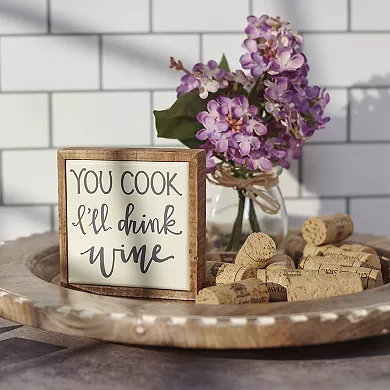 A decorative wooden sign with the phrase \“YOU COOK I'll drink wine\“ is displayed alongside a bunch of corks and a vase with lilac flowers.