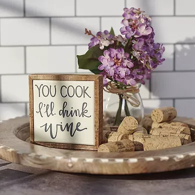 A decorative wooden sign with the phrase \“YOU COOK I'll drink wine\“ is displayed alongside a bunch of corks and a vase with lilac flowers.