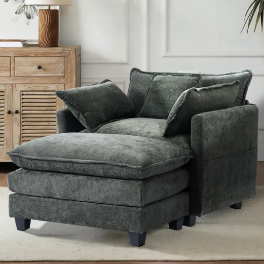 A plush gray chaise lounge with multiple soft pillows, situated on a light-colored rug next to a wooden cabinet with shutter-style doors and a table lamp.