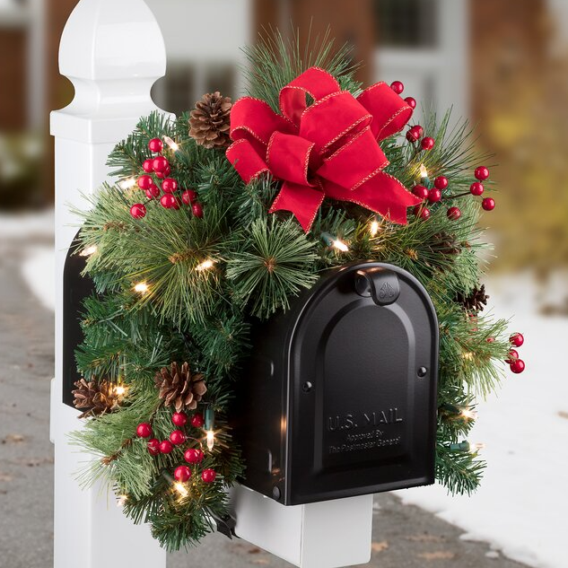 A festive mailbox swag adorned with pine branches, red berries, pinecones, a large red ribbon, and twinkling lights, creating a holiday-themed decoration for a mailbox.