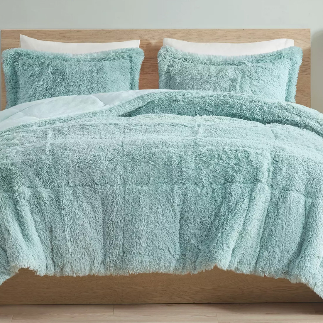 A bed with a fluffy light teal comforter set, including two matching pillow shams, against a wooden headboard.