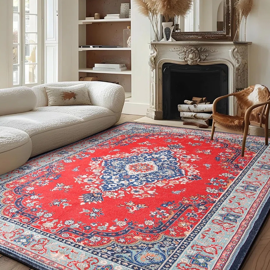 A living room with a red and blue patterned rug, a white upholstered sectional sofa, and a decorative fireplace with logs. A light wooden chair with a fur cushion is also present.