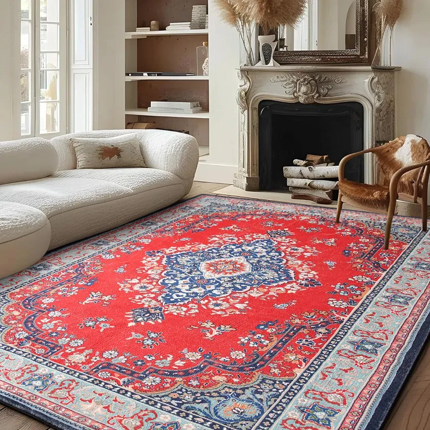 A living room with a red and blue patterned rug, a white upholstered sectional sofa, and a decorative fireplace with logs. A light wooden chair with a fur cushion is also present.