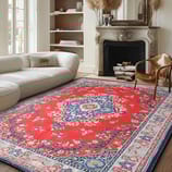 A living room with a red and blue patterned rug, a white upholstered sectional sofa, and a decorative fireplace with logs. A light wooden chair with a fur cushion is also present.