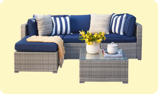 Outdoor patio furniture set with a gray wicker base, featuring dark blue cushions and a variety of pillows. Includes an L-shaped sectional sofa and a matching glass-top coffee table, accented with a vase of yellow flowers and a decorative throw.