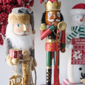 A nutcracker figurine is dressed in a woodland theme, featuring a gray herringbone coat, fur-lined hat, beard, and holding a tiny wrapped gift and tree.