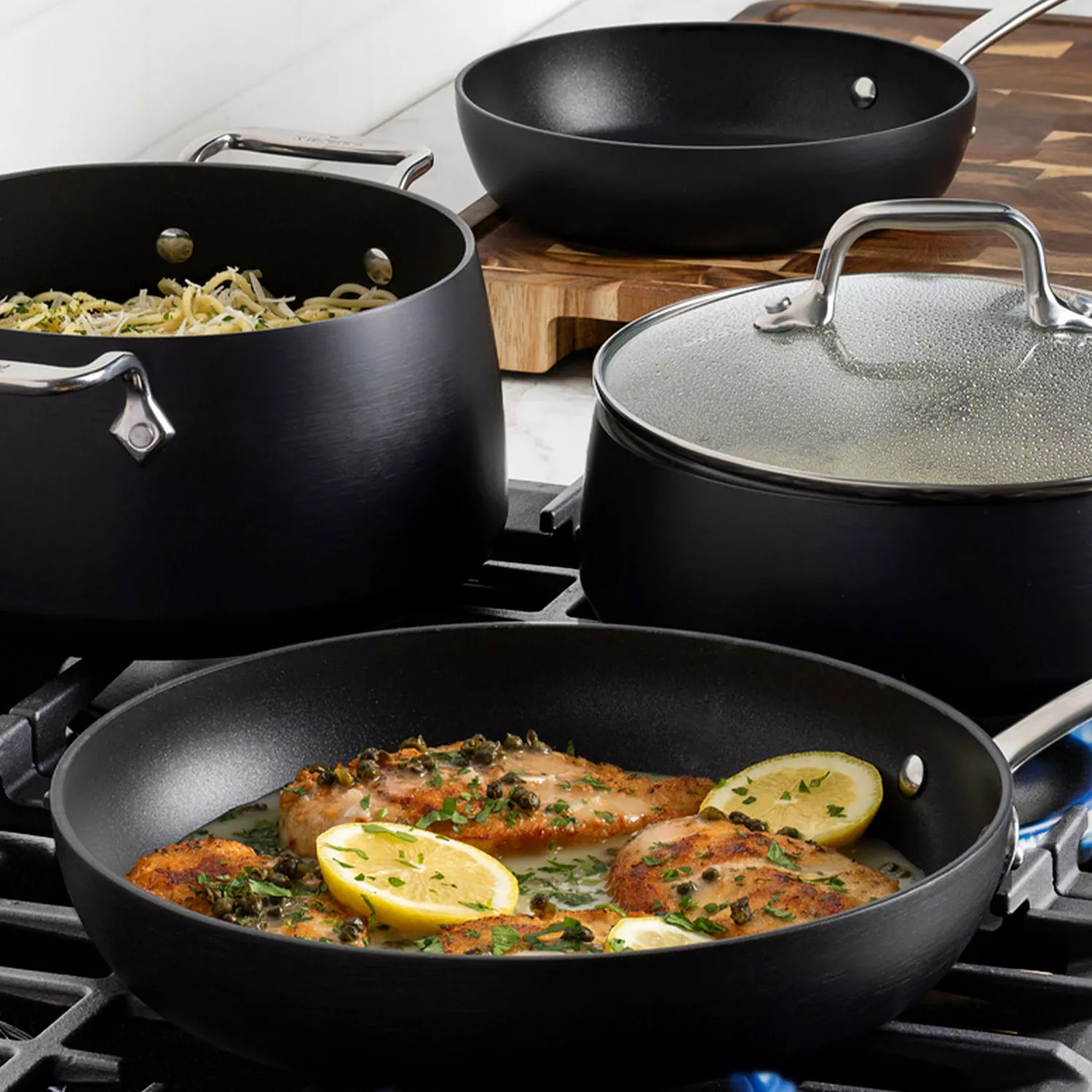 Black nonstick cookware set on a stove, featuring a frying pan with chicken and lemon slices, a pot with pasta, a lidded saucepan, and a skillet on a wooden board.