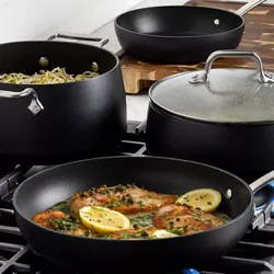 Black nonstick cookware set on a stove, featuring a frying pan with chicken and lemon slices, a pot with pasta, a lidded saucepan, and a skillet on a wooden board.