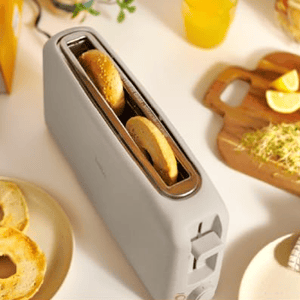 A toaster with a long, wide slot fitting two slices side-by-side, surrounded by toasted bagels and lemons on a cutting board.