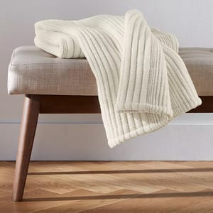 A cream-colored, ribbed throw blanket drapes over a beige fabric bench with wooden legs on a wooden floor.
