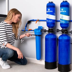 A woman is kneeling next to an Aquasana water filtration system, adjusting the blue filter component. The system includes multiple blue canisters for filtering and conditioning water.