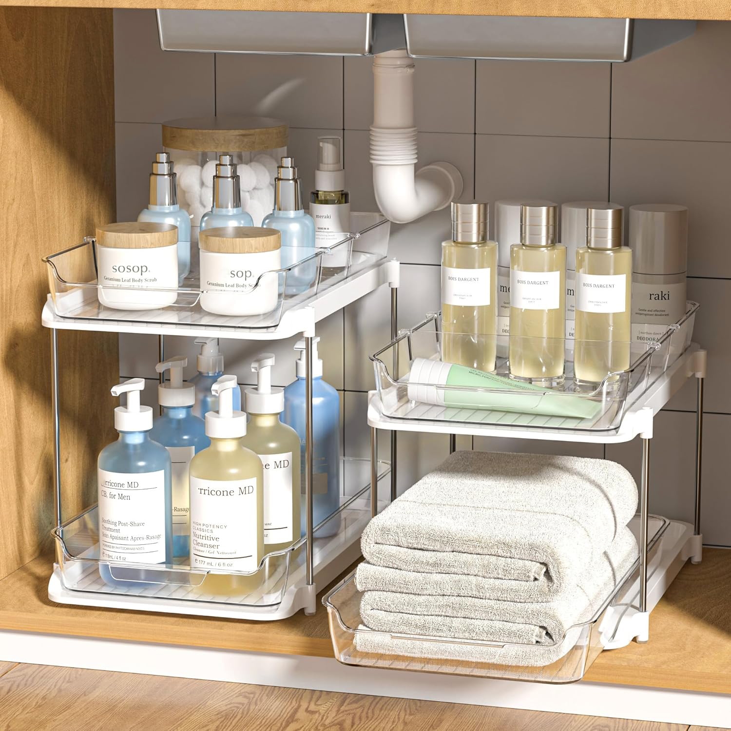 The 2pk Cabinet Organizers feature two-tier storage with metal wire baskets, holding various toiletries and folded towels, designed to fit under a sink. They have a compact design ideal for maximizing space in small areas.