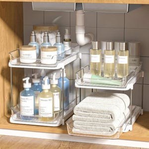 The 2pk Cabinet Organizers feature two-tier storage with metal wire baskets, holding various toiletries and folded towels, designed to fit under a sink. They have a compact design ideal for maximizing space in small areas.