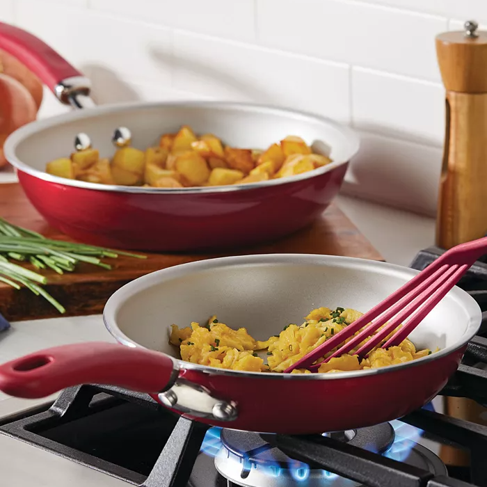 Rachael Ray Nonstick Fry Pans in red, featuring two sizes: one cooking scrambled eggs and the other with potatoes. Both have rubberized handles and are suitable for stove-top use with visible flame underneath.