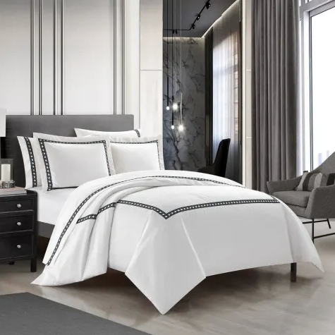 A modern bedroom features a neatly made bed with white bedding accented by black trim, set against a backdrop of luxurious decor including a gray chair, dark nightstand, and elegant lighting.