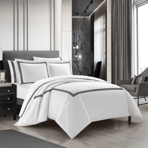A modern bedroom features a neatly made bed with white bedding accented by black trim, set against a backdrop of luxurious decor including a gray chair, dark nightstand, and elegant lighting.